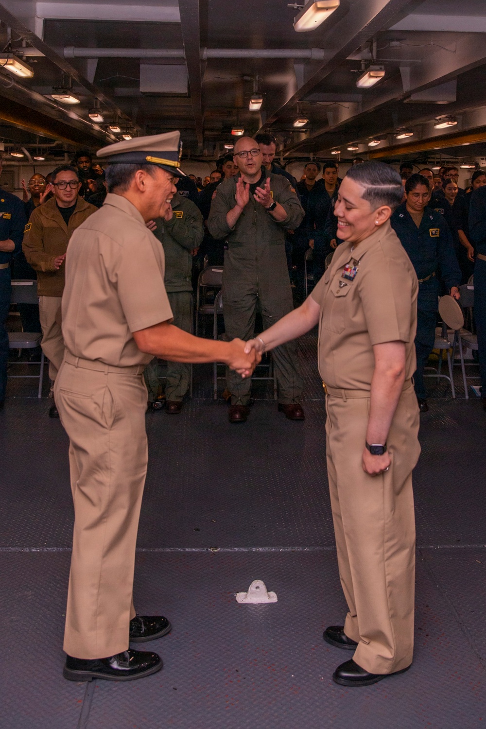 Two U.S. Navy personnel in Service Khaki uniforms are shaking hands during what appears to be a formal military ceremony aboard a naval vessel. The officer on the left is wearing a combination cover (cap) and appears to be of senior rank, while the individual on the right displays ribbons/medals on their uniform. A crowd of sailors and at least one person in a flight suit are applauding in the background.