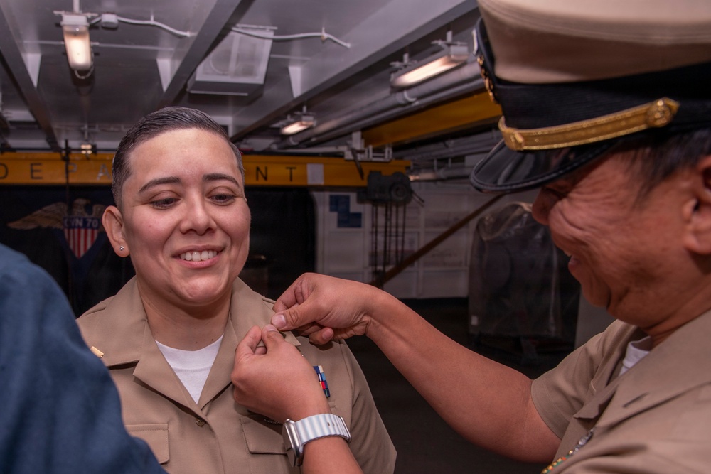 A U.S. Navy ceremony is taking place aboard what appears to be a naval vessel, where a senior officer is pinning a medal or insignia onto a junior sailor's uniform. The recipient is smiling broadly, suggesting this is a positive recognition or promotion event. Partial text 'CVN 70' is visible in the background, indicating this may be aboard USS Carl Vinson.