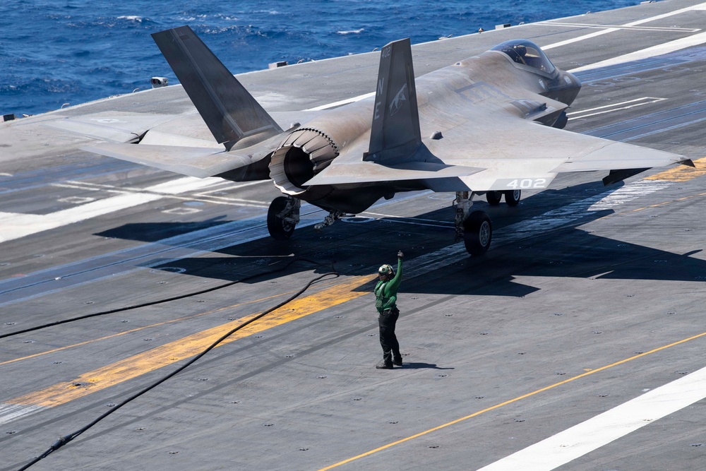 A carrier deck operations scene showing an F-35C Lightning II stealth fighter jet (bureau number 402) positioned on an aircraft carrier flight deck. A flight deck crew member wearing a green jersey and helmet is giving hand signals in front of the aircraft. The ocean is visible in the background, confirming an at-sea operational environment.