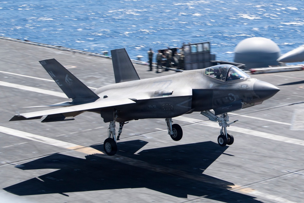 An F-35B Lightning II stealth fighter jet is captured landing or taking off on the flight deck of a naval amphibious assault ship. The aircraft's landing gear is fully deployed and in contact with or very close to the deck surface. Several personnel are visible in the background near ship superstructure elements, with open ocean visible beyond the deck.