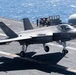 An F-35B Lightning II stealth fighter jet is captured landing or taking off on the flight deck of a naval amphibious assault ship. The aircraft's landing gear is fully deployed and in contact with or very close to the deck surface. Several personnel are visible in the background near ship superstructure elements, with open ocean visible beyond the deck.
