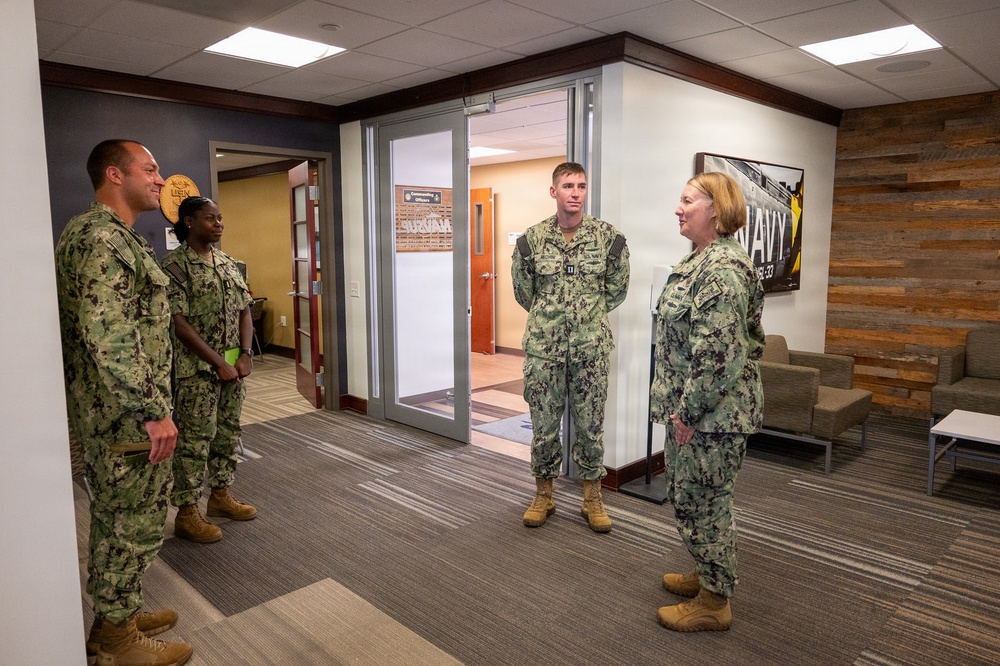 Four U.S. Navy personnel in NWU Type III (woodland digital camouflage) uniforms are gathered in what appears to be a modern military facility lobby or reception area. A senior female officer on the right appears to be speaking with or briefing the other three service members. The setting includes a 'NAVY' branded wall display and a rustic wood accent wall, suggesting a recruiting or administrative building.