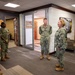 Four U.S. Navy personnel in NWU Type III (woodland digital camouflage) uniforms are gathered in what appears to be a modern military facility lobby or reception area. A senior female officer on the right appears to be speaking with or briefing the other three service members. The setting includes a 'NAVY' branded wall display and a rustic wood accent wall, suggesting a recruiting or administrative building.
