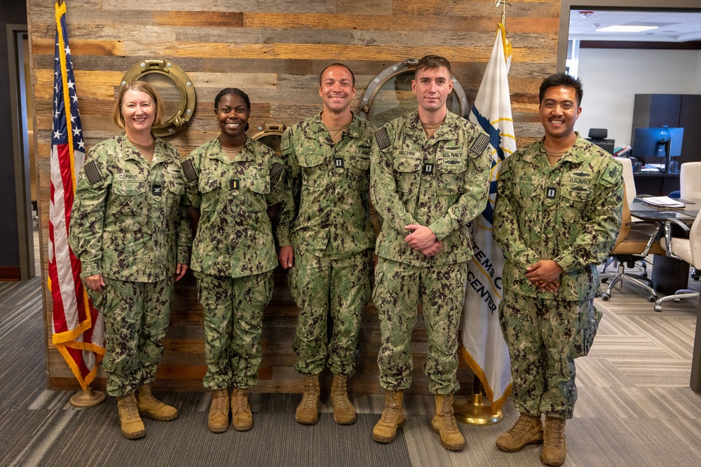 Five U.S. Navy personnel in NWU Type III (digital woodland camouflage) uniforms pose for a group photo in what appears to be a military office or command center. The group includes both male and female sailors of varying ranks, standing in front of a decorative reclaimed wood wall with brass porthole accents. An American flag and a Navy command flag are visible flanking the group.