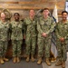 Five U.S. Navy personnel in NWU Type III (digital woodland camouflage) uniforms pose for a group photo in what appears to be a military office or command center. The group includes both male and female sailors of varying ranks, standing in front of a decorative reclaimed wood wall with brass porthole accents. An American flag and a Navy command flag are visible flanking the group.