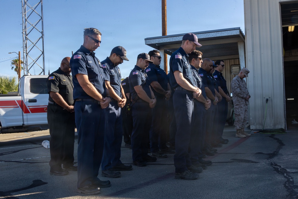 A group of uniformed first responders, appearing to be firefighters or emergency services personnel, stand in a line with heads bowed and hands clasped in a moment of silence or prayer. The setting appears to be outside a fire station or emergency services facility. One individual on the far right is wearing military camouflage uniform, suggesting a multi-agency or ceremonial gathering.