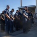 A group of uniformed first responders, appearing to be firefighters or emergency services personnel, stand in a line with heads bowed and hands clasped in a moment of silence or prayer. The setting appears to be outside a fire station or emergency services facility. One individual on the far right is wearing military camouflage uniform, suggesting a multi-agency or ceremonial gathering.