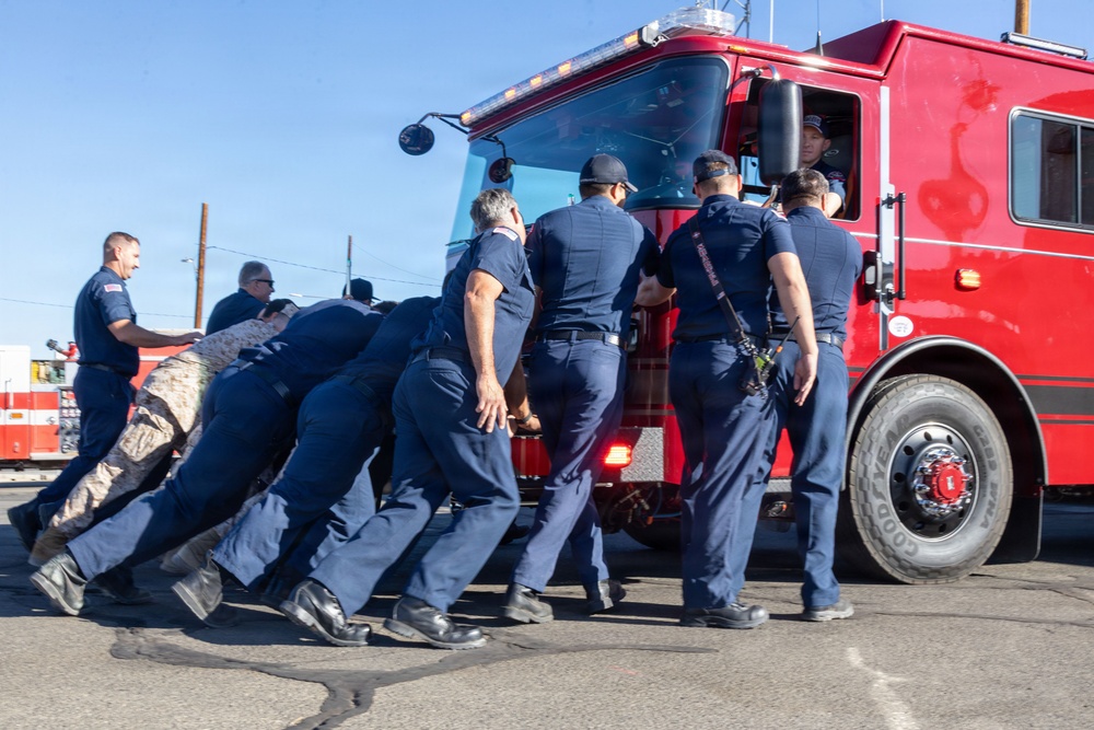 A group of approximately eight to nine firefighters dressed in navy blue uniforms are collectively pushing a large red fire truck from behind on a paved surface. One person in military camouflage pants is also participating in the push. A driver is visible inside the cab of the fire truck, and the activity appears to be a strength challenge or training exercise conducted outdoors under clear blue skies.