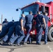 A group of approximately eight to nine firefighters dressed in navy blue uniforms are collectively pushing a large red fire truck from behind on a paved surface. One person in military camouflage pants is also participating in the push. A driver is visible inside the cab of the fire truck, and the activity appears to be a strength challenge or training exercise conducted outdoors under clear blue skies.