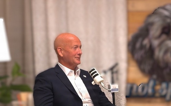 A bald man in a navy blazer and white pants sits in a light wooden armchair, appearing to be a guest on a podcast or interview show. He has a microphone clipped to his lapel and is seated in what appears to be a styled studio set. Behind him is a wooden wall with a painted dog portrait and partial text reading 'not special'.
