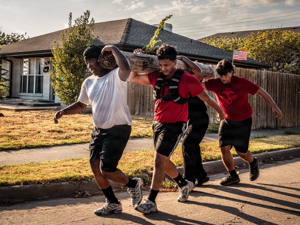 Three young men are carrying a large wooden log on their shoulders while running or walking briskly along a residential street. They appear to be engaged in a physical training exercise, likely a team workout or athletic conditioning drill. The setting is a suburban neighborhood with houses, trees, and an American flag visible in the background.