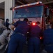 A group of individuals in military camouflage uniforms and navy blue firefighter uniforms are manually pushing a red fire truck into or out of a fire station bay. The fire truck has visible emergency lights and partial text reading 'COMBAT' on the front. One firefighter in navy blue stands to the right observing while others physically push the vehicle.