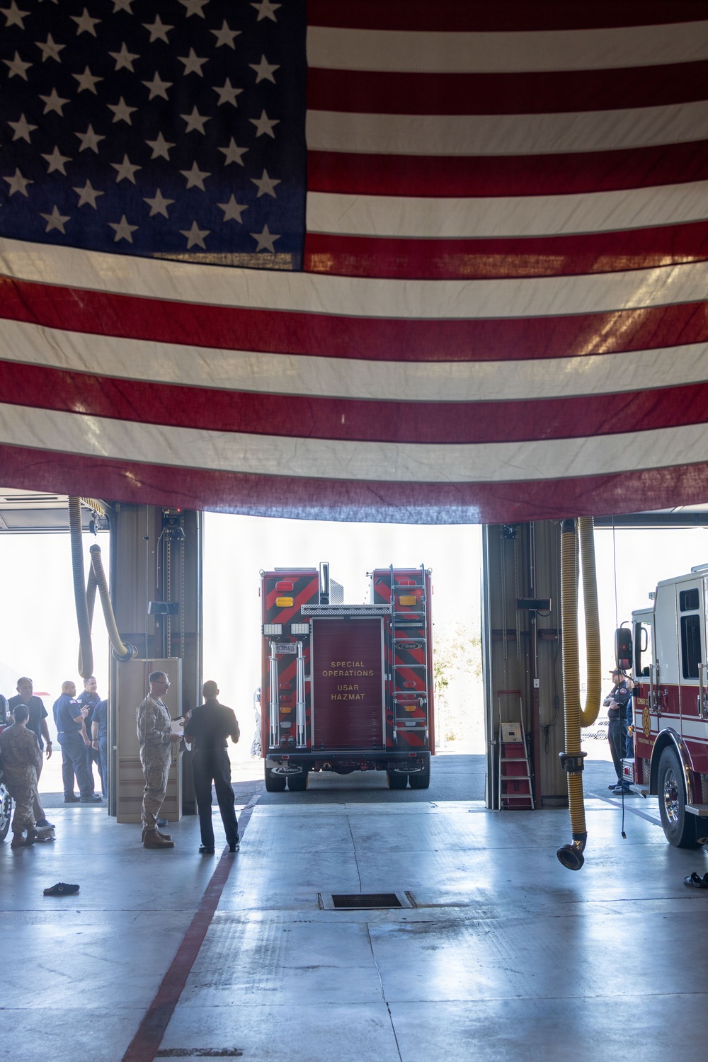 The image shows the interior of a fire station bay with a large American flag draped across the overhead door opening. A red Special Operations USAR/HAZMAT vehicle is visible backing out or parked at the bay entrance, while several personnel — some in military uniforms and others in civilian attire — are gathered inside. The scene appears to be a formal event or ceremony, suggested by the prominent flag display and the mixed group of attendees.