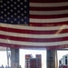 The image shows the interior of a fire station bay with a large American flag draped across the overhead door opening. A red Special Operations USAR/HAZMAT vehicle is visible backing out or parked at the bay entrance, while several personnel — some in military uniforms and others in civilian attire — are gathered inside. The scene appears to be a formal event or ceremony, suggested by the prominent flag display and the mixed group of attendees.