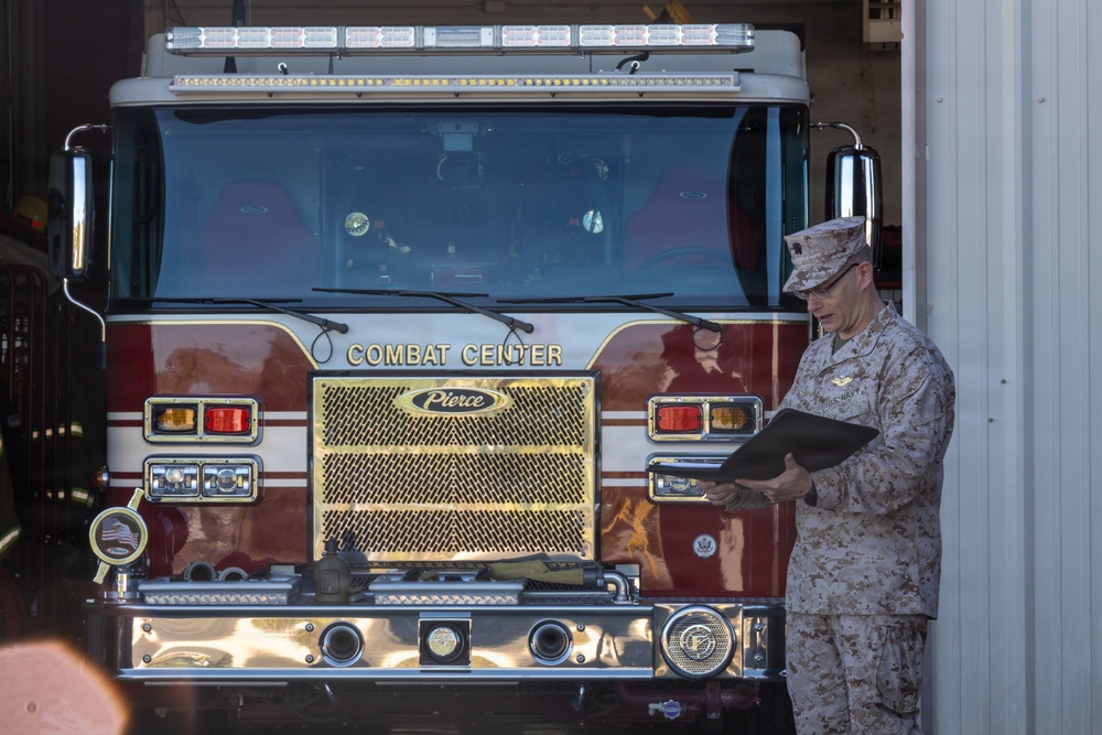 A military officer in Marine Corps Combat Utility Uniform (MCCUU) stands beside a red Pierce fire truck labeled 'Combat Center,' reading from a dark folder or clipboard. The setting appears to be a fire station bay at a military installation, likely a Marine Corps Combat Center. The scene suggests an official inspection, ceremony, or administrative review.