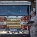 A military officer in Marine Corps Combat Utility Uniform (MCCUU) stands beside a red Pierce fire truck labeled 'Combat Center,' reading from a dark folder or clipboard. The setting appears to be a fire station bay at a military installation, likely a Marine Corps Combat Center. The scene suggests an official inspection, ceremony, or administrative review.