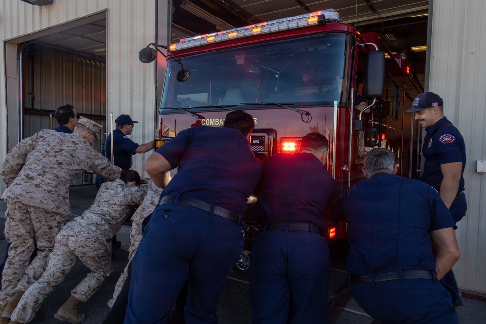 A group of individuals in military camouflage uniforms and navy blue firefighter uniforms are manually pushing a red fire truck into or out of a fire station bay. The fire truck has amber and red emergency lights visible and partial text reading 'COMBAT' on the front. One firefighter in navy blue stands to the right observing while others physically push the vehicle.