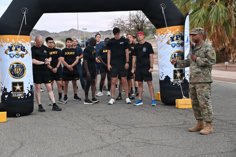 A U.S. Army event at Yuma Proving Ground (YPG) showing a group of soldiers and personnel in PT (physical training) uniforms gathered under an inflatable arch at what appears to be a race or fitness event start/finish line. A soldier in full camouflage uniform (ACUs) stands to the right, appearing to address or brief the group. The setting is an outdoor paved area with desert mountains visible in the background.