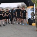 A U.S. Army event at Yuma Proving Ground (YPG) showing a group of soldiers and personnel in PT (physical training) uniforms gathered under an inflatable arch at what appears to be a race or fitness event start/finish line. A soldier in full camouflage uniform (ACUs) stands to the right, appearing to address or brief the group. The setting is an outdoor paved area with desert mountains visible in the background.