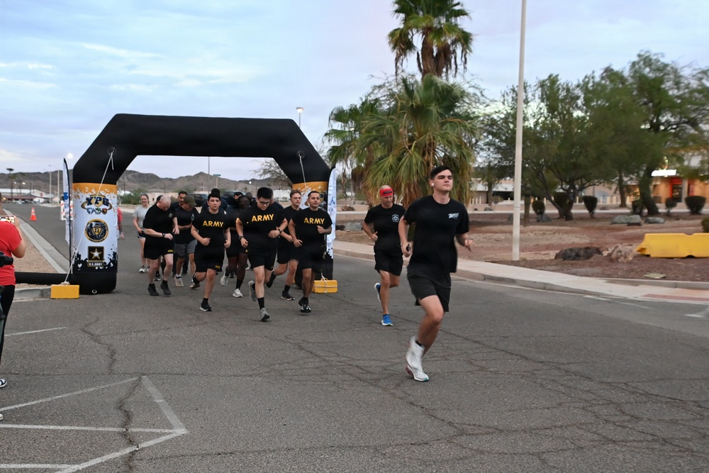 A group of U.S. Army personnel and civilians are participating in a running event, passing under a large black inflatable arch at what appears to be a military installation. Several participants are wearing black Army PT (Physical Training) shirts with gold 'ARMY' lettering. The setting appears to be a desert environment, likely in the American Southwest, based on the palm trees, arid landscape, and mountain backdrop.