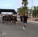 A group of U.S. Army personnel and civilians are participating in a running event, passing under a large black inflatable arch at what appears to be a military installation. Several participants are wearing black Army PT (Physical Training) shirts with gold 'ARMY' lettering. The setting appears to be a desert environment, likely in the American Southwest, based on the palm trees, arid landscape, and mountain backdrop.