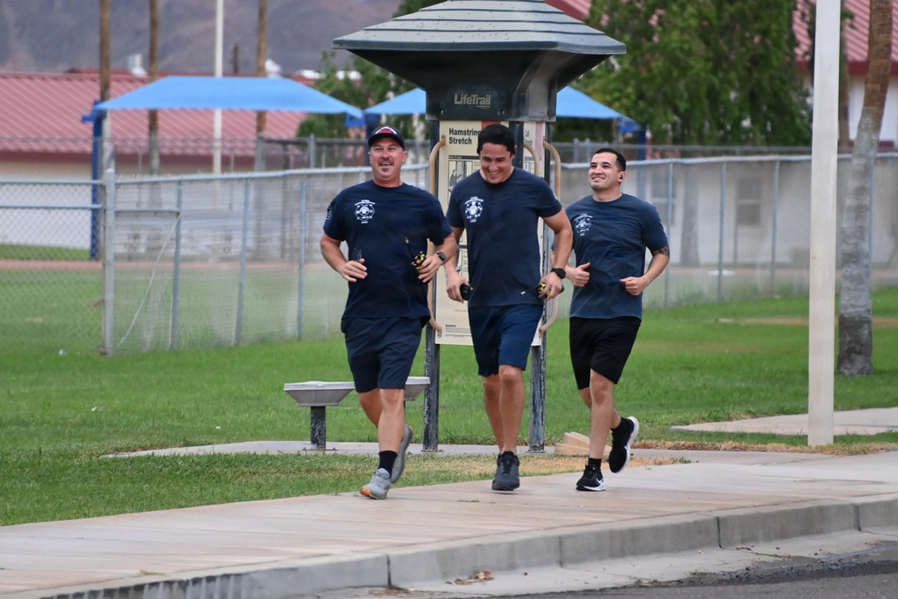 Three men in matching navy blue t-shirts and shorts are jogging together along a paved path in what appears to be a park or recreational facility. All three are smiling and appear to be in good spirits during their run. A 'LifeTrail' fitness station sign for 'Hamstring Stretch' is visible in the background.