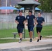 Three men in matching navy blue t-shirts and shorts are jogging together along a paved path in what appears to be a park or recreational facility. All three are smiling and appear to be in good spirits during their run. A 'LifeTrail' fitness station sign for 'Hamstring Stretch' is visible in the background.