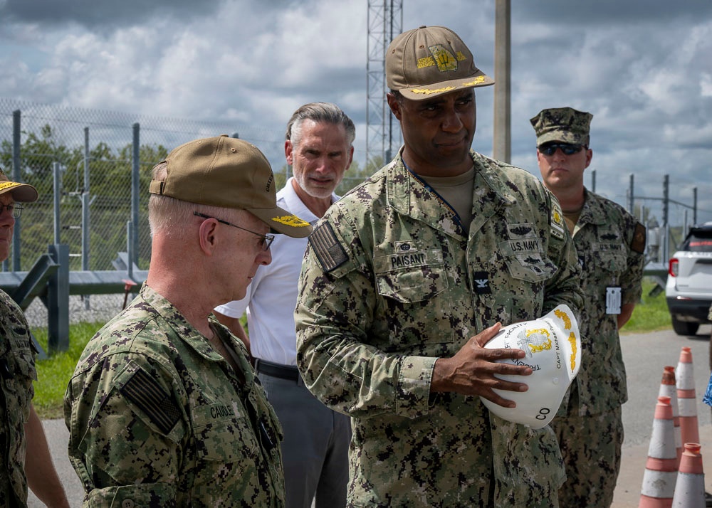 Two U.S. Navy officers in NWU Type III (digital camouflage) uniforms are engaged in a discussion, with one officer named 'PAISANT' holding a white hard hat inscribed with text including 'CAPT' and what appears to be a facility or command name. Several other military and civilian personnel are visible in the background. The setting appears to be an outdoor military installation or construction/industrial site, indicated by the chain-link fencing, traffic cones, and hard hat.