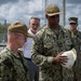 Two U.S. Navy officers in NWU Type III (digital camouflage) uniforms are engaged in a discussion, with one officer named 'PAISANT' holding a white hard hat inscribed with text including 'CAPT' and what appears to be a facility or command name. Several other military and civilian personnel are visible in the background. The setting appears to be an outdoor military installation or construction/industrial site, indicated by the chain-link fencing, traffic cones, and hard hat.