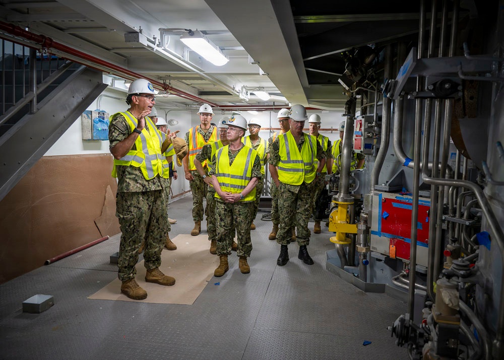 A group of military personnel in U.S. Navy camouflage uniforms are conducting an inspection or tour of an industrial or shipboard facility. All individuals are wearing high-visibility yellow safety vests and white hard hats, suggesting an active construction or maintenance environment. One individual on the left appears to be briefing or explaining something to the group near complex piping and mechanical equipment.