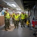 A group of military personnel in U.S. Navy camouflage uniforms are conducting an inspection or tour of an industrial or shipboard facility. All individuals are wearing high-visibility yellow safety vests and white hard hats, suggesting an active construction or maintenance environment. One individual on the left appears to be briefing or explaining something to the group near complex piping and mechanical equipment.