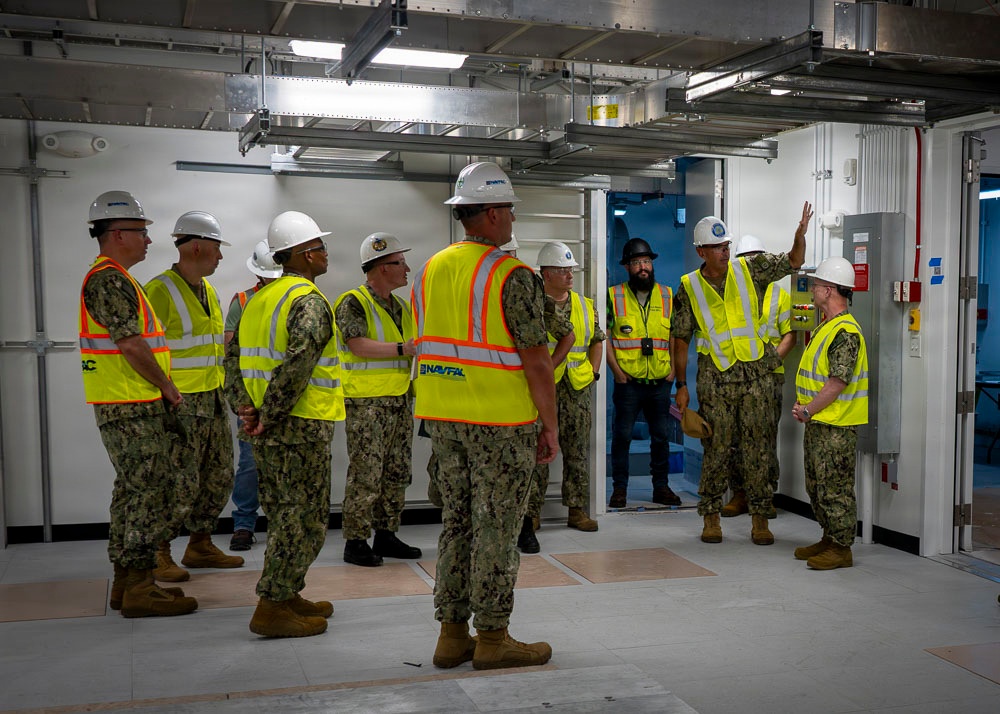 A group of U.S. Navy personnel in camouflage uniforms are gathered inside what appears to be a newly constructed or renovated facility for a briefing or walkthrough. All individuals are wearing high-visibility yellow safety vests and white hard hats, consistent with a construction site safety inspection or facility tour. One individual on the right side appears to be gesturing and speaking to the group, suggesting a leadership or instructional role.