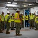 A group of U.S. Navy personnel in camouflage uniforms are gathered inside what appears to be a newly constructed or renovated facility for a briefing or walkthrough. All individuals are wearing high-visibility yellow safety vests and white hard hats, consistent with a construction site safety inspection or facility tour. One individual on the right side appears to be gesturing and speaking to the group, suggesting a leadership or instructional role.