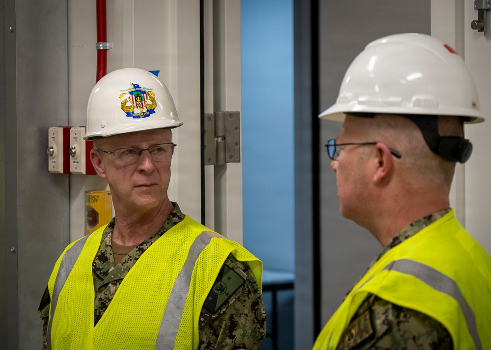 Two individuals wearing U.S. Navy digital camouflage uniforms and high-visibility safety vests are engaged in conversation inside what appears to be a facility under construction or inspection. Both wear white hard hats and glasses; the person on the left has a military insignia emblem on their helmet. The setting suggests an official safety inspection or facility tour.