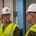 Two individuals wearing U.S. Navy digital camouflage uniforms and high-visibility safety vests are engaged in conversation inside what appears to be a facility under construction or inspection. Both wear white hard hats and glasses; the person on the left has a military insignia emblem on their helmet. The setting suggests an official safety inspection or facility tour.
