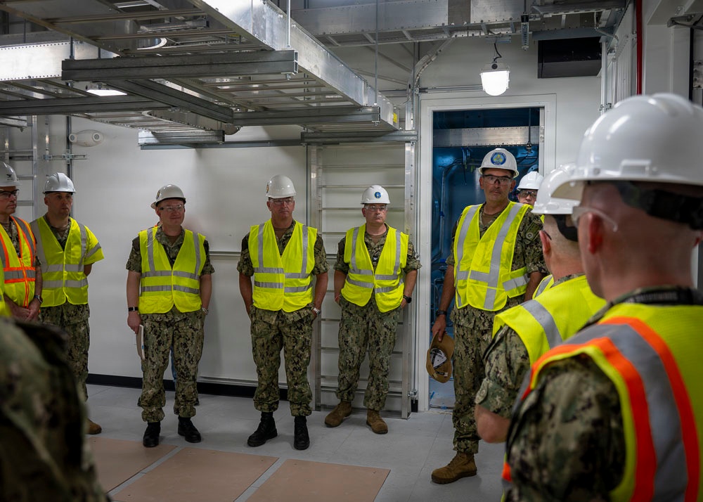 A group of military personnel in U.S. Navy camouflage uniforms are gathered inside what appears to be a facility under construction or inspection. All individuals are wearing high-visibility yellow safety vests and white hard hats, indicating a safety-regulated environment. One person facing away from the camera appears to be briefing or addressing the group.