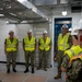 A group of military personnel in U.S. Navy camouflage uniforms are gathered inside what appears to be a facility under construction or inspection. All individuals are wearing high-visibility yellow safety vests and white hard hats, indicating a safety-regulated environment. One person facing away from the camera appears to be briefing or addressing the group.