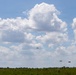 The image shows a military airborne operation with multiple paratroopers descending over a flat, open grassy field under a partly cloudy blue sky. Approximately six to seven parachutes are visible at various altitudes, suggesting a mass tactical jump. The scene appears to be a training exercise or operational jump onto a drop zone.