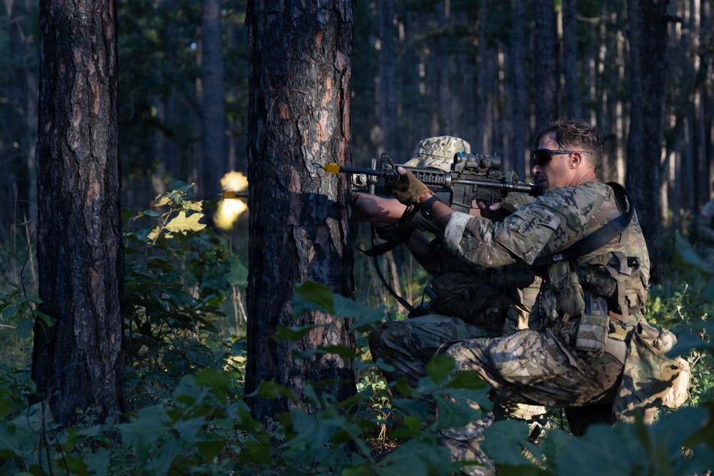 A soldier in multicam/OCP camouflage uniform is positioned in a kneeling stance behind a tree in a forested environment, actively firing a rifle. A muzzle flash is visible at the barrel of the weapon, indicating live or blank fire. A second soldier wearing a boonie hat is partially visible behind the tree.