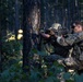 A soldier in multicam/OCP camouflage uniform is positioned in a kneeling stance behind a tree in a forested environment, actively firing a rifle. A muzzle flash is visible at the barrel of the weapon, indicating live or blank fire. A second soldier wearing a boonie hat is partially visible behind the tree.