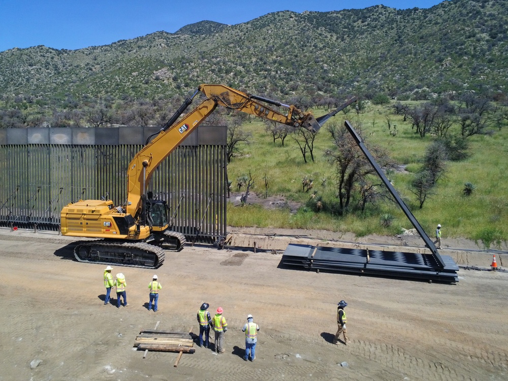 An aerial view shows a large yellow Caterpillar (CAT) excavator being used in the construction or installation of a border wall with tall vertical steel bollards. Several construction workers wearing high-visibility vests and hard hats are observing and coordinating the operation. The site is located in a semi-arid, mountainous landscape with green vegetation visible in the background.