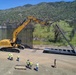 An aerial view shows a large yellow Caterpillar (CAT) excavator being used in the construction or installation of a border wall with tall vertical steel bollards. Several construction workers wearing high-visibility vests and hard hats are observing and coordinating the operation. The site is located in a semi-arid, mountainous landscape with green vegetation visible in the background.