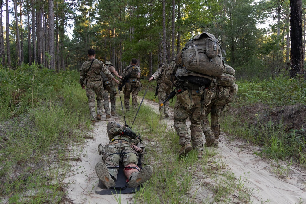 A group of military personnel in camouflage uniforms are conducting a field training exercise along a sandy forest trail. One soldier is being dragged on a litter or drag device along the ground, simulating a casualty evacuation scenario. The group is moving through a pine forest environment with heavy rucksacks and tactical gear.