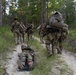 A group of military personnel in camouflage uniforms are conducting a field training exercise along a sandy forest trail. One soldier is being dragged on a litter or drag device along the ground, simulating a casualty evacuation scenario. The group is moving through a pine forest environment with heavy rucksacks and tactical gear.