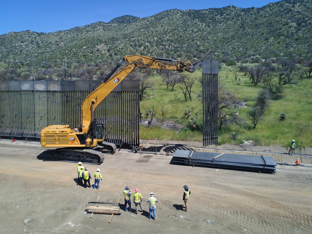 An aerial view shows a large CAT excavator actively installing or working on tall steel bollard barrier panels along a border fence line. Several construction workers wearing high-visibility safety vests and hard hats are gathered nearby, with stacked steel panels on the ground awaiting installation. The scene is set against a backdrop of green, vegetation-covered mountains under a clear blue sky.