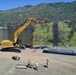 An aerial view shows a large CAT excavator actively installing or working on tall steel bollard barrier panels along a border fence line. Several construction workers wearing high-visibility safety vests and hard hats are gathered nearby, with stacked steel panels on the ground awaiting installation. The scene is set against a backdrop of green, vegetation-covered mountains under a clear blue sky.