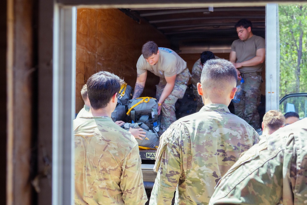 A group of military personnel in camouflage uniforms are loading or unloading equipment from the back of a large box truck or trailer. Several soldiers stand outside observing while others inside the truck handle gear including backpacks and other military equipment. The scene appears to take place outdoors near a wooded area during daylight hours.