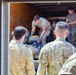A group of military personnel in camouflage uniforms are loading or unloading equipment from the back of a large box truck or trailer. Several soldiers stand outside observing while others inside the truck handle gear including backpacks and other military equipment. The scene appears to take place outdoors near a wooded area during daylight hours.