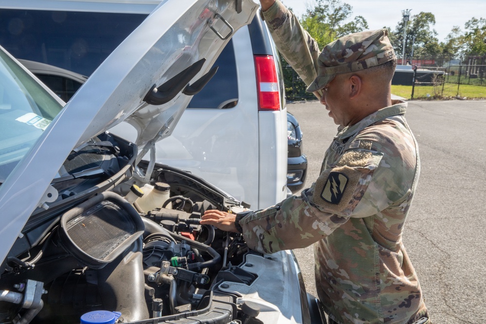 A U.S. Army soldier in Operational Camouflage Pattern (OCP) uniform is inspecting or performing maintenance on the engine of a white van or large vehicle. The soldier is leaning over the open hood, examining components in the engine bay. The setting appears to be a paved parking area on what looks like a military installation.