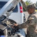 A U.S. Army soldier in Operational Camouflage Pattern (OCP) uniform is inspecting or performing maintenance on the engine of a white van or large vehicle. The soldier is leaning over the open hood, examining components in the engine bay. The setting appears to be a paved parking area on what looks like a military installation.